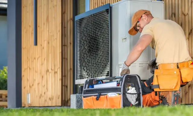 Instalator w pasie narzędziowym sprawdza jednostkę zewnętrzną pompy ciepła przy drewnianym domu.