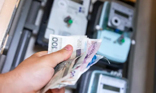 Ręka trzyma polskie banknoty przed licznikami energii. Zastanawiasz się, jak zmienić dostawcę prądu, by oszczędzić?