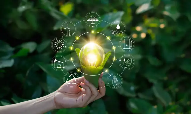 Dłoń trzyma żarówkę z zielonymi liśćmi, otoczoną ikonami symbolizującymi niekonwencjonalne źródła energii: wiatr, słońce, wodę, biomasę i paliwa.