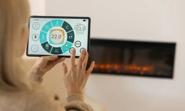 Kobieta steruje inteligentnym domem przez tablet, kontrolując temperaturę i inne funkcje. Widoczny jest ekran z ikonami i wskaźnikami, a w tle kominek.