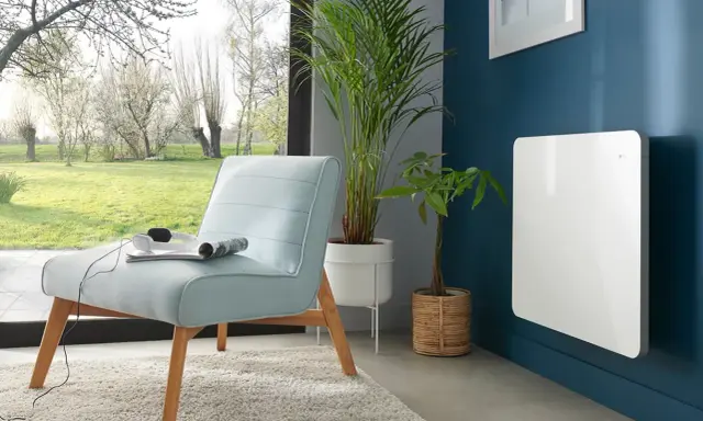 Nowoczesny, biały grzejnik elektryczny na granatowej ścianie. Idealny jako jaki grzejnik do pokoju 10m2, obok jasnoniebieskiej kanapy i roślin.
