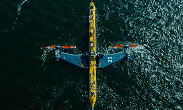 Podwodna turbina wykorzystująca energię pływów, z charakterystycznymi żółto-niebieskimi elementami i pomarańczowymi śmigłami, zanurzona w ciemnych wodach oceanu.