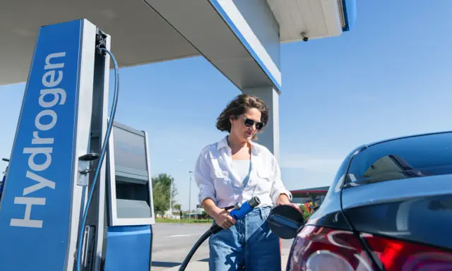 Kobieta tankuje auta na wodór. Stacja z napisem "Hydrogen" i niebieska dystrybutor.