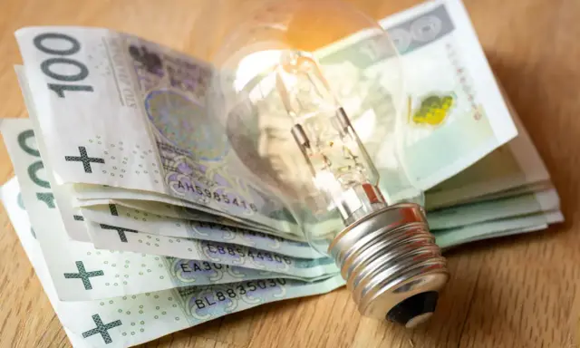 Żarówka na stosie banknotów 100 zł symbolizuje wysokie koszty energii i potrzebę dopłat do prądu.