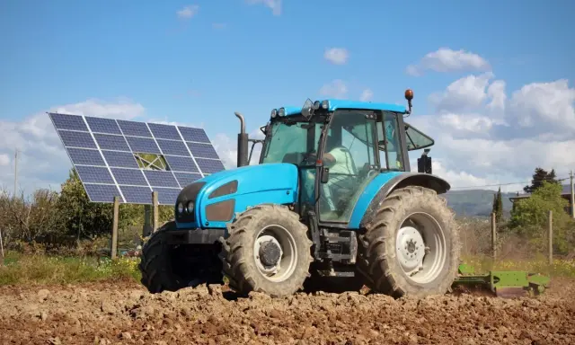 Błękitny traktor pracuje na polu, obok paneli fotowoltaika arimr. Słońce i chmury na niebie.