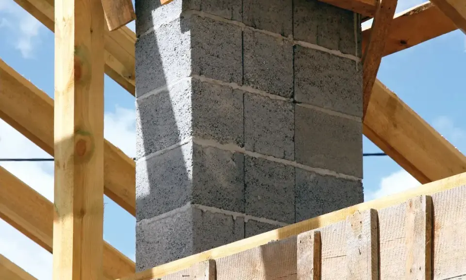 Budowa komina systemowego z bloczków betonowych, gotowego do podłączenia pieca. Drewniana konstrukcja dachu w tle.