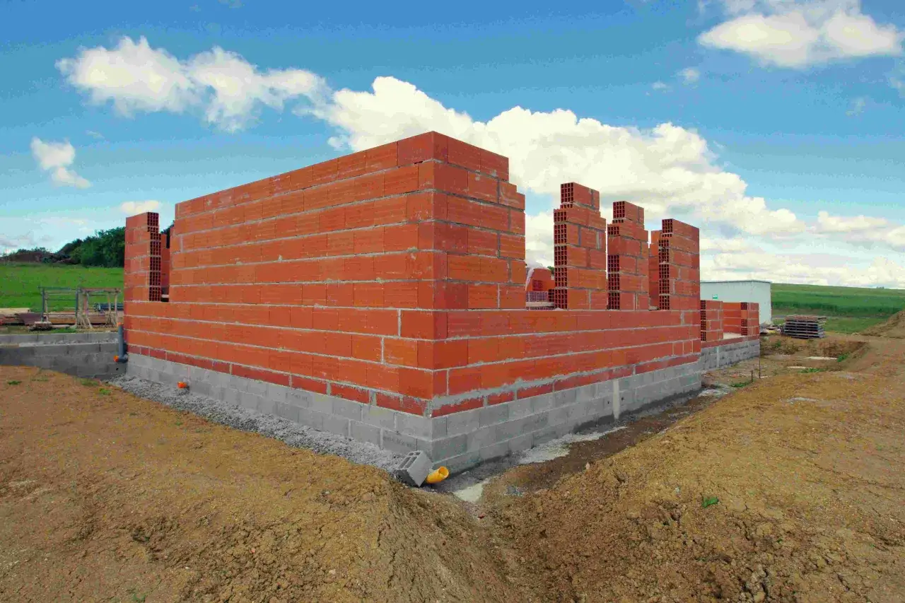 Budowa domu w toku: czerwone ściany z pustaków piętrzą się na betonowym fundamencie. W tle zielone wzgórza i błękitne niebo z chmurami. Zastanawiasz się, ile trwa budowa domu?