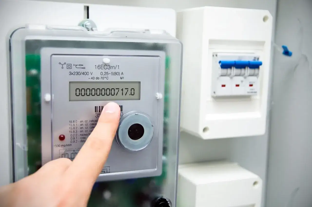 Palec wskazuje na cyfrowy licznik energii elektrycznej, odczytując jego symbole i wskazania. Obok widoczny jest wyłącznik nadprądowy.